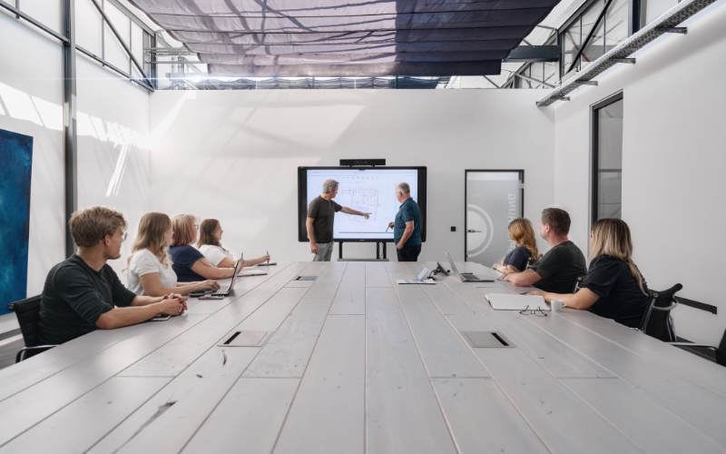 Zwei Männer präsentieren an einem großen Touchscreen in einem modernen Konferenzraum, sechs Personen sitzen an einem langen Tisch und folgen aufmerksam der Besprechung.