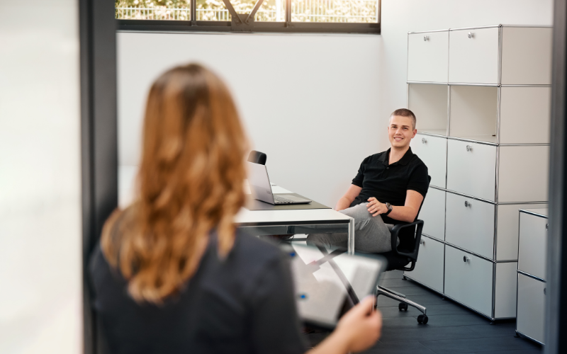 Junge Frau mit Tablet betritt ein Büro, in dem ein junger Mann entspannt am Schreibtisch sitzt und lächelt – lockere Gesprächssituation in einem modernen Arbeitsumfeld.