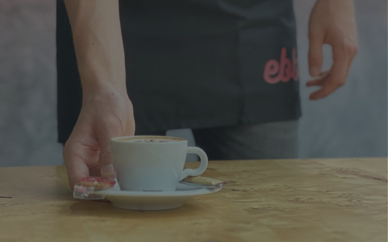 Ein Servicemitarbeiter im Ebbtron stellt eine frisch zubereitete Kaffeespezialität mit Gebäck auf den Tisch.