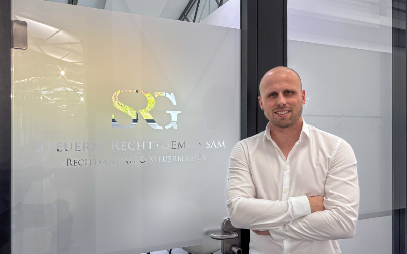 Steuerberater Nick Rohrbeck steht freundlich schauend vor einer Glastür mit dem Logo seiner Firma